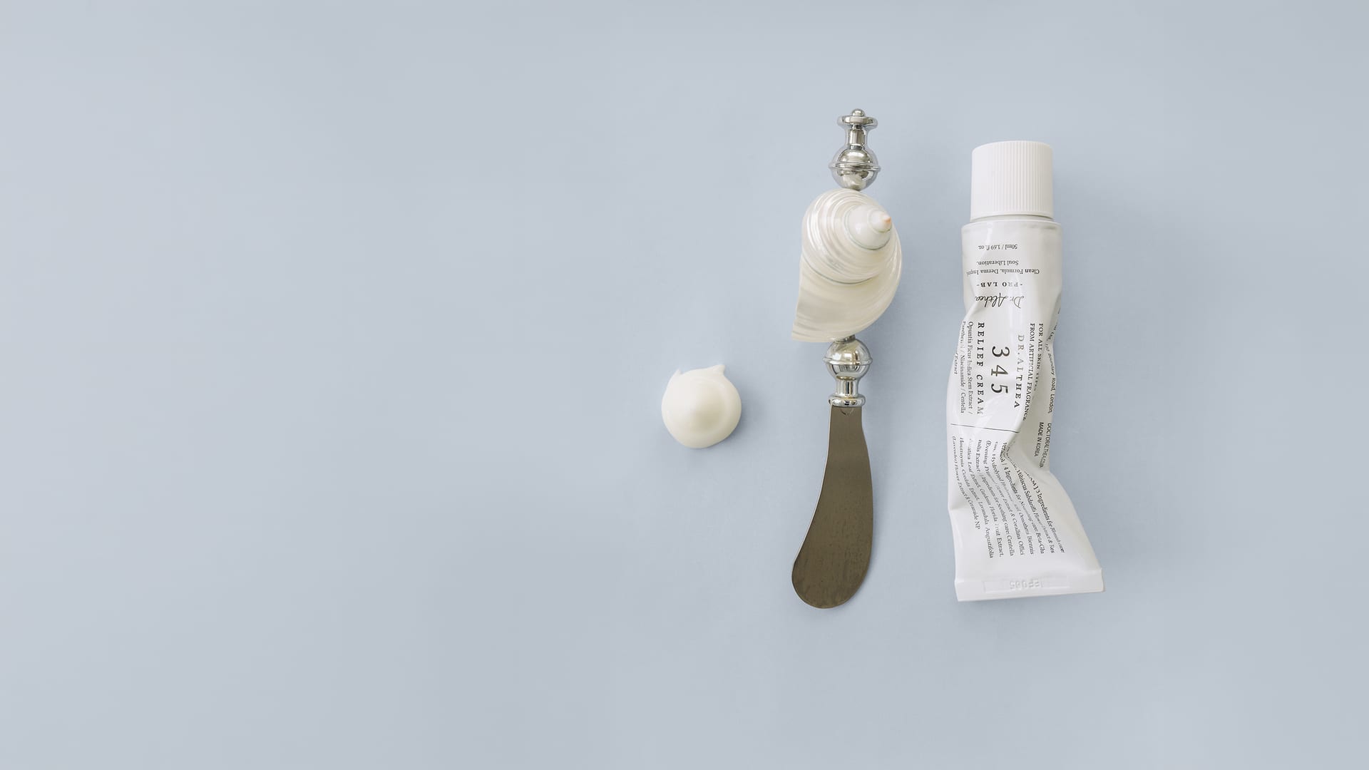 Cremetube, Spatel und eine Muschel mit einem Klecks Creme auf hellem Blaugrau